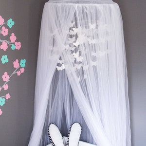 Pode incluir: Mosquiteiro branco com dossel e um m&oacute;bile de borboletas brancas pendurado dentro. O mosquiteiro est&aacute; drapeado sobre um ber&ccedil;o branco com um coelho branco e um travesseiro em forma de estrela dourada.