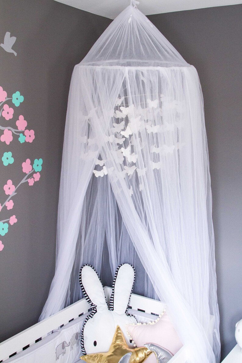 Pode incluir: Mosquiteiro branco com dossel e um m&oacute;bile de borboletas brancas pendurado dentro. O mosquiteiro est&aacute; drapeado sobre um ber&ccedil;o branco com um coelho branco e um travesseiro em forma de estrela dourada.