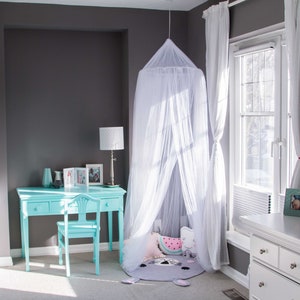 Pode incluir: Uma cama com dossel branco com um tapete estampado em cinza e rosa e uma almofada de melancia rosa e verde. A cama est&aacute; em um quarto com uma escrivaninha e uma cadeira brancas, uma estante de livros e uma janela com cortinas brancas.