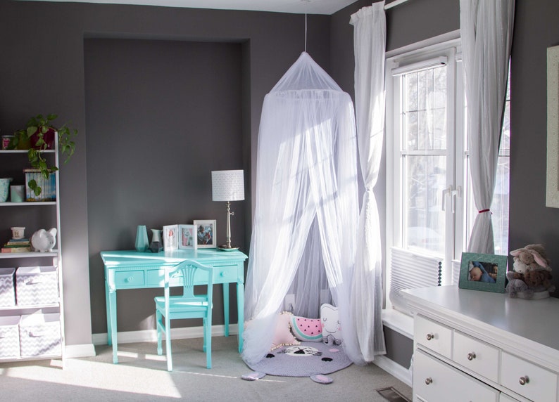 Pode incluir: Uma cama com dossel branco com um tapete estampado em cinza e rosa e uma almofada de melancia rosa e verde. A cama est&aacute; em um quarto com uma escrivaninha e uma cadeira brancas, uma estante de livros e uma janela com cortinas brancas.