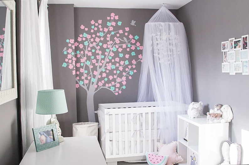Pode incluir: Um ber&ccedil;o branco com um dossel branco e um decalque floral rosa e verde na parede atr&aacute;s dele. H&aacute; uma c&ocirc;moda branca com uma l&acirc;mpada branca e uma foto emoldurada no topo.