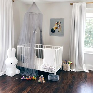 Pode incluir: Um ber&ccedil;o branco com um dossel cinza e um cobertor branco com pompons. H&aacute; uma luz noturna de coelho branco e um trem de brinquedo de madeira no ch&atilde;o. O quarto tem cortinas brancas e um piso de madeira marrom.