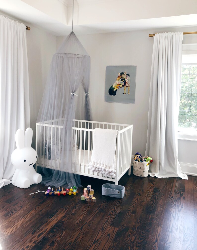 Pode incluir: Um ber&ccedil;o branco com um dossel cinza e um cobertor branco com pompons. H&aacute; uma luz noturna de coelho branco e um trem de brinquedo de madeira no ch&atilde;o. O quarto tem cortinas brancas e um piso de madeira marrom.