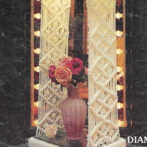 May include: A white macrame lampshade with a white fur trim hangs from the ceiling. The lampshade is in front of a mirror with a row of light bulbs on each side. A vase with pink and white roses sits on a table below the lampshade.