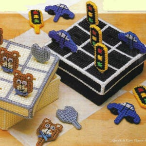 May include: Two plastic canvas boxes with a game board design. One box has a black and white game board with traffic lights and cars. The other box has a white and blue game board with bears and hearts.  The boxes are made with plastic canvas and are decorated with colorful yarn.