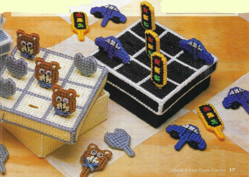 May include: Two plastic canvas boxes with a game board design. One box has a black and white game board with traffic lights and cars. The other box has a white and blue game board with bears and hearts.  The boxes are made with plastic canvas and are decorated with colorful yarn.