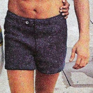 May include: A man wearing blue swim trunks with a button closure. He is standing with his feet bare on a concrete surface.