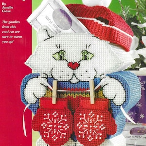 Puede incluir: Un gato blanco de crochet sosteniendo dos guantes rojos con diseños de copos de nieve. El gato lleva un gorro rojo y blanco de punto con un pompón blanco. El gato sostiene un paquete de mezcla de bebida de chocolate Rich Cocoa. El texto "Cocoa Kitty" está en la parte superior de la imagen.