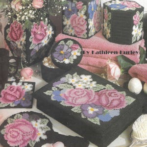 May include: A set of black canvas craft projects featuring floral designs with roses, pansies, and daisies. The set includes a tissue box cover, a small box, a round box, a square coaster, a hand mirror, and a rectangular box. The text "By Kathleen Hurley" is visible in the image.
