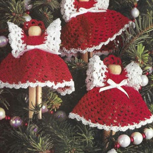 May include: Three crocheted angel ornaments with white wings and red dresses. The angels are hanging on a Christmas tree.