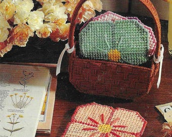 Puede incluir: Un juego de cuatro posavasos de ganchillo con forma de flor en una cesta de mimbre marrón. Los posavasos son en tonos de rosa, verde y amarillo.