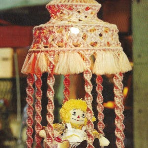 May include: A handmade, crochet dollhouse-style hanging decoration. The structure is primarily peach and cream-colored yarn, with red accents. A doll with yellow hair and a white dress sits inside, holding a small gift. The piece has a whimsical, vintage aesthetic.
