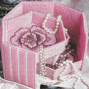 Könnte beinhalten: Eine rosa, sechseckige, dreistufige Schmuckschatulle mit einem Blumendesign im Inneren. Die Schatulle besteht aus einem gewebten Material und hat ein gestreiftes Muster auf der Außenseite. Perlenketten sind über die Schatulle drapiert.