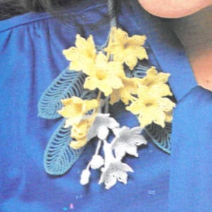 May include: A crocheted flower brooch with yellow and white flowers and blue leaves. The brooch is pinned to a blue shirt.