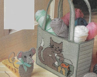 Op de afbeelding: Een grijze plastic deurstopper in de vorm van een muis die een stuk kaas vasthoudt. De deurstopper ligt naast een grijze plastic draagtas met een kattenontwerp en de woorden "Mouse & Cheese DOORSTOP". De draagtas is gevuld met bollen garen in verschillende kleuren.