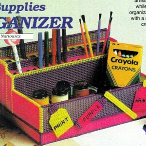 May include: A colourful plastic organiser for art supplies, with compartments for pens, pencils, paint, and crayons. The organiser has a purple and pink base with yellow accents and labels that say "Print" and "Purple". The box is labelled "Art Supplies Organiser" and "Crayola Crayons".