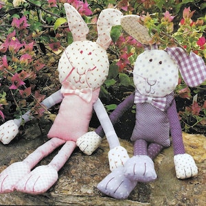May include: Two handmade fabric bunny toys with long limbs and bow ties. The bunny on the left is pink with white polka dots and the bunny on the right is purple with white polka dots.