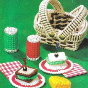 May include: A miniature plastic canvas picnic basket with a red and white gingham tablecloth, two cans, a sandwich, a slice of cheese, and a cupcake.