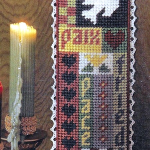May include: A white crocheted bookmark with a colorful cross-stitch design featuring the words "Peace", "Faith", and "Hope". The bookmark has a brown tassel.