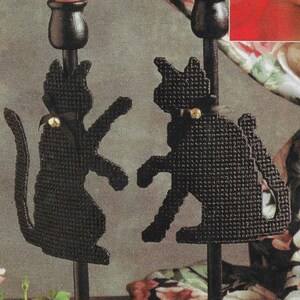 May include: Two black cat candle holders with a black ribbon and a gold bell on each. The candle holders are made of plastic canvas and are attached to black metal candle sticks.