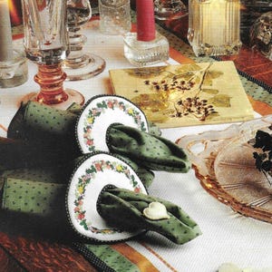 May include: A white tablecloth with a green and black trim, two green napkin rings with floral embroidery, and two white napkins. The table is set with candles and glasses.