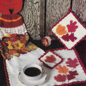 May include: A five-piece crochet dining set with an autumn theme. The set includes a leaf-shaped pot holder, a towel, a napkin ring, a coaster, and a placemat. The set features red, orange, and yellow leaf designs.