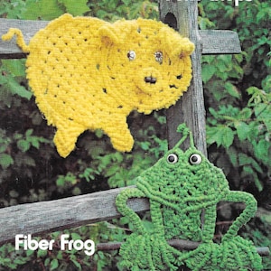 May include: A yellow crocheted pig with black eyes and a pink nose, and a green crocheted frog with black eyes, both hanging on a wooden fence. The pig is labeled "Pork Chops" and the frog is labeled "Fiber Frog".