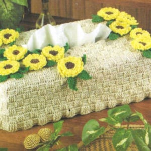 Puede incluir: Una funda para caja de pañuelos de tejido crochet con un diseño tejido y un motivo de girasol. La funda está decorada con girasoles de crochet amarillos y hojas verdes.