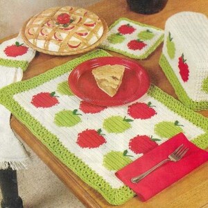 May include: A set of crocheted apple-themed kitchen accessories, including a table runner, placemats, a bread warmer, and a potholder. The accessories are white with green and red apples.