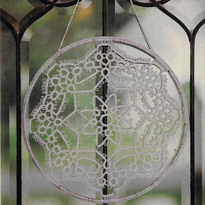 Könnte beinhalten: Ein kreisförmiges, weißes Spitzendeckchen, das an einer dünnen Schnur aufgehängt ist, wird vor einem Fenster gezeigt. Das filigrane Spitzendesign weist florale und geometrische Muster auf. Das Deckchen wird von einem dünnen, hellfarbigen Ring umrahmt.