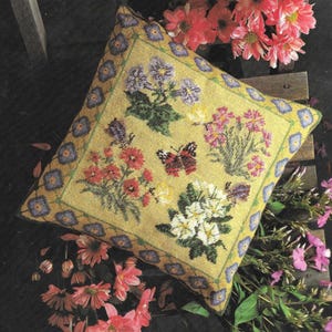 Könnte beinhalten: Ein quadratisches gelbes Kissen mit einem blauen Rand und einem Blumendesign. Das Kissen zeigt vier verschiedene Arten von Blumen und Schmetterlingen in Rot-, Rosa-, Weiß- und Grüntönen.