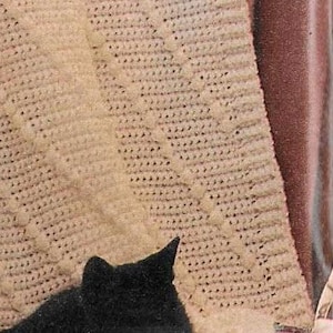 Puede incluir: Un gato negro está durmiendo en una manta blanca de ganchillo con un patrón de diamante. La manta está drapeada sobre una silla con un cojín de estampado floral.