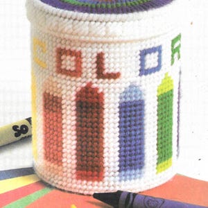 May include: A white canvas container with a rainbow design on the lid and the word "COLOR" on the front. The container is filled with crayons.