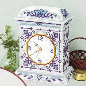 Puede incluir: Un reloj blanco y morado con un diseño floral. La esfera del reloj tiene números romanos y un borde dorado.