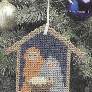 May include: A brown and tan plastic ornament shaped like a house with a blue background. Inside the house are three figures: a woman in orange, a man in gray, and a baby in white. The ornament has a white ribbon attached to the top for hanging.