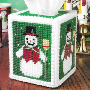 May include: A white tissue box cover with a green and white cross-stitch design. The design features a snowman wearing a black top hat, red vest, and holding a broom. The tissue box has a white border and a green top.