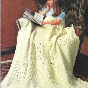 May include: A young girl sits in a chair reading a book while wrapped in a white crocheted blanket with a cable stitch pattern. The blanket has a delicate, lacy look.