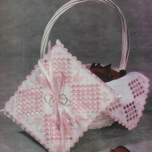 May include: A white and pink embroidered ring pillow with a pink satin ribbon and two gold rings. The pillow is in a white wicker basket with rose petals.