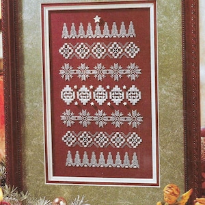 Puede incluir: Un sampler de punto de cruz enmarcado con un fondo rojo y bordado blanco. El sampler presenta una variedad de diseños de temática invernal, incluyendo copos de nieve, árboles y estrellas. El marco es marrón con un borde dorado.