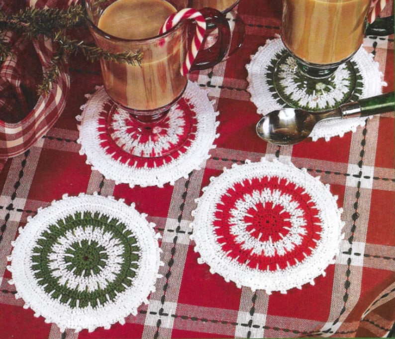 May include: Four crocheted coasters in red, white, and green. The coasters are circular and have a center design with a white border. The coasters are on a red and white checkered tablecloth.