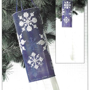 May include: A blue and white plastic canvas ornament with a snowflake design. The ornament is hanging from a pine tree branch.