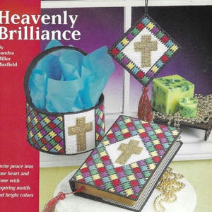 May include: A craft project pattern book titled "Heavenly Brilliance" by Sandra Miller Maxfield. The book cover features a colorful cross design with a gold cross. The book is next to a colorful cross-shaped box with a gold cross. The book and box are surrounded by gold beads and a green candle.