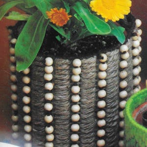 May include: A cylindrical planter with a woven, rope-like texture, adorned with vertical rows of small, light-colored beads. The planter holds a vibrant plant with green leaves and bright yellow and orange flowers, creating a natural, decorative piece.