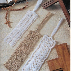 May include: Three crocheted bookmarks with fringe tassels. Two are white and one is brown. The bookmarks are laid out on a white surface with a wooden cross and a rosary.