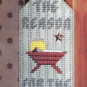 Puede incluir: Un colgador de puerta de crochet blanco y azul con el texto "The Reason for the Season" y una representación de la escena de la natividad con una estrella amarilla y un pesebre rojo.