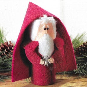 Puede incluir: Un adorno de Santa Claus de fieltro rojo con barba blanca y una estrella plateada. El adorno lleva una túnica roja y un gorro rojo puntiagudo.