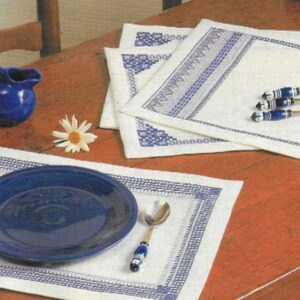 May include: A dining table setting featuring white placemats with blue embroidered borders. A blue pitcher, plates, and spoons with blue and white handles are arranged on the table. Artificial daisies add a decorative touch to the scene.