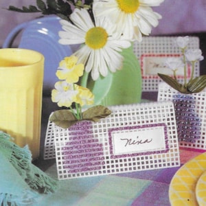 Peut inclure: Cartes de place en plastique blanc avec des bordures perlées et des noms écrits à l'encre noire. Les cartes de place sont décorées de fleurs et sont disposées sur une table avec un verre jaune, un vase bleu et une nappe à fleurs.