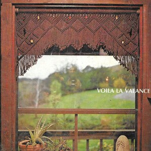 May include: A brown macrame valance with a diamond pattern and fringe hangs in a window. The valance is decorated with small, round beads. The window looks out onto a green, wooded landscape.