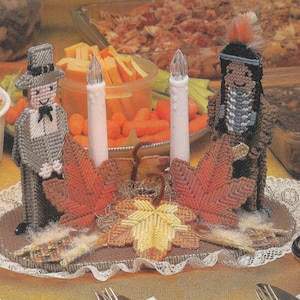 Pode incluir: Um centro de mesa temático de Ação de Graças com duas figuras, uma com traje de peregrino e a outra com vestimenta nativa americana, ladeando duas velas brancas. As figuras são cercadas por folhas de outono e uma base marrom.